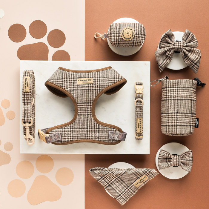 Plaid bow tie, harness, leash, collar, and pouches set in brown tones on marble and brown surface.