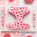 A flat lay photo of the Pupberry Fields Adjustable Neck Harness, Lead & Collar Bundle by Cocopup London, showcasing a pink harness set adorned with a strawberry pattern. The set includes an adjustable neck harness, leash, and collar, all featuring matching designs. The items are beautifully arranged on a white surface with strawberry-themed paper accents surrounding them, making it the perfect ensemble for stylish dog walks.