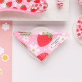 A pink dog bandana with a stylish strawberry and white flower pattern, named "Pupberry Fields" from Cocopup London, is laid out on a white surface. The bandana is accompanied by other matching dog accessories such as a harness and bow tie, perfect for the fashionable pup.