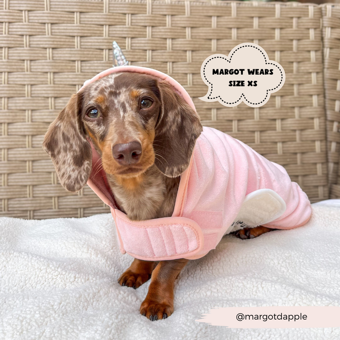 A small brown and tan Dachshund sporting a pink, unicorn-inspired hoodie with a horn on the hood lounges on a cushioned surface. A speech bubble reads "Margot wears size XS." The Instagram handle "@margotdapple" appears at the bottom. She dreams of the PRE-ORDER Pupicorn Drying Robe by Cocopup London, crafted from premium microfibre.