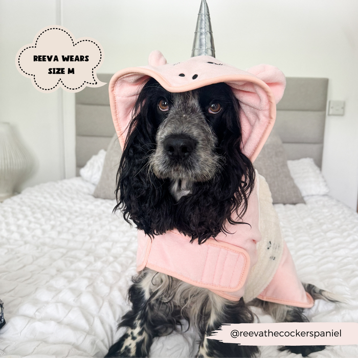 A black and white cocker spaniel is sitting on a bed, dressed in a pink PRE-ORDER Pupicorn Drying Robe with a unicorn horn on the hood from Cocopup London. Text on the image reads, "REEVA WEARS SIZE M." The Instagram handle "@reevathecockerspaniel" is in the bottom-right corner.