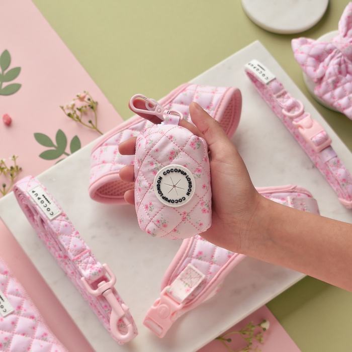 Hand holds a pink quilted floral poop bag holder with matching dog accessories in the background.