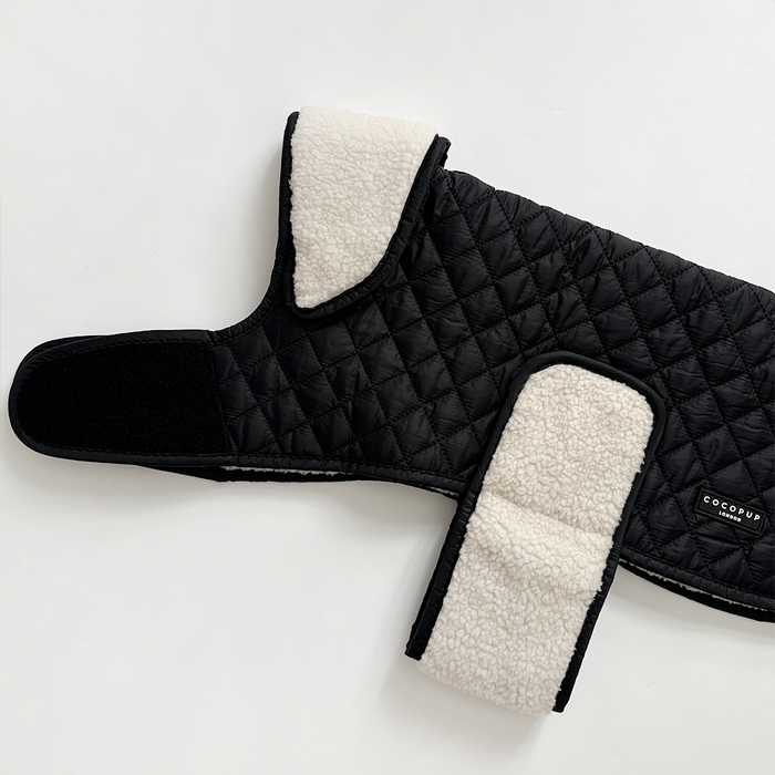Black quilted dog coat with white teddy lining and Velcro straps, displayed flat on a white background.