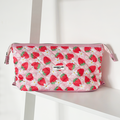Pink quilted makeup bag with strawberry and floral pattern on a white shelf with strawberries and flowers nearby.
