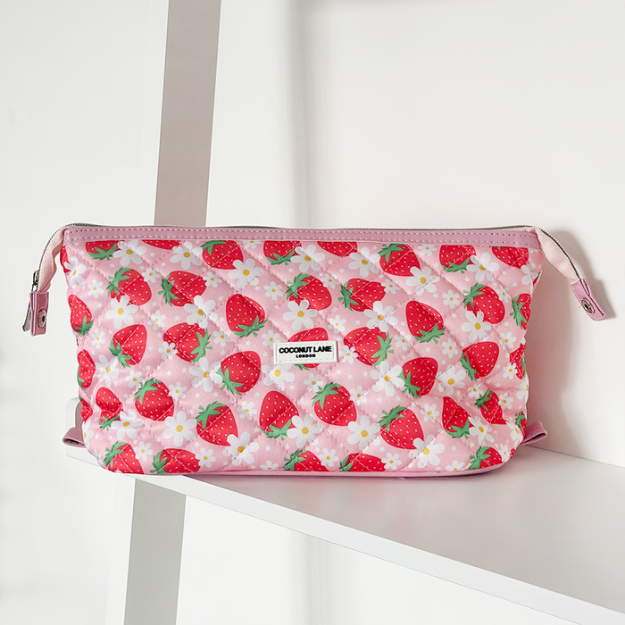 Pink quilted makeup bag with strawberry and floral pattern on a white shelf with strawberries and flowers nearby.