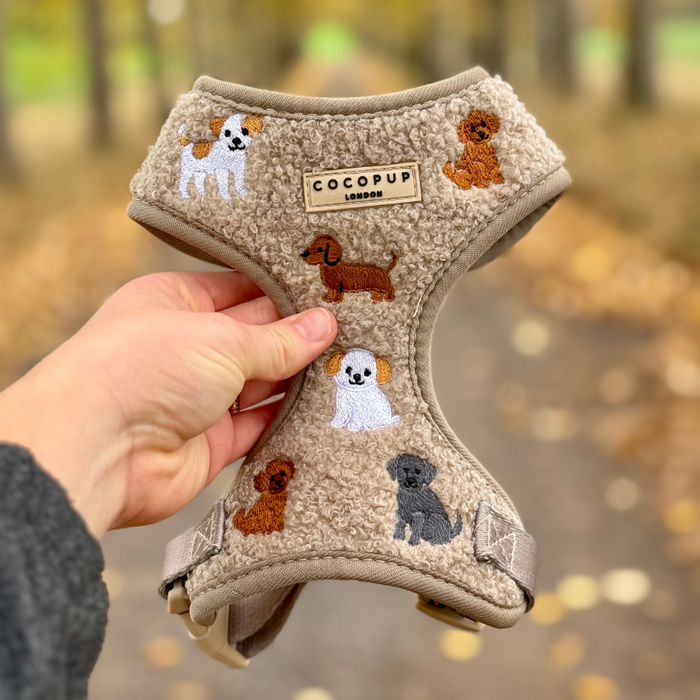 Hand holding a brown teddy fleece dog harness with adjustable neck, shown outdoors on a leafy autumn path.