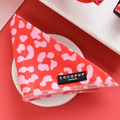 A neatly folded Red & Pink Leopard Bandana labeled "Cocopup London" rests on a white plate. The background showcases a red surface with a partially visible white and black patterned object in the corner, perfectly complementing the Cocopup range of matching products.