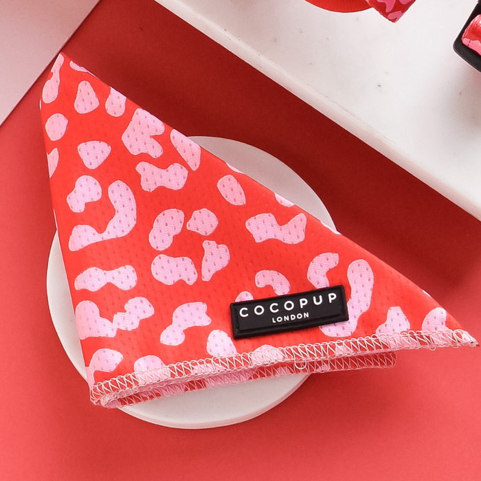 A neatly folded Red & Pink Leopard Bandana labeled "Cocopup London" rests on a white plate. The background showcases a red surface with a partially visible white and black patterned object in the corner, perfectly complementing the Cocopup range of matching products.