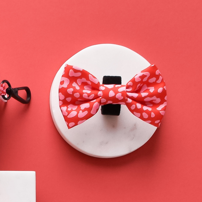 Red and pink leopard print bow tie with pink hearts, displayed on a white platform against a red background.