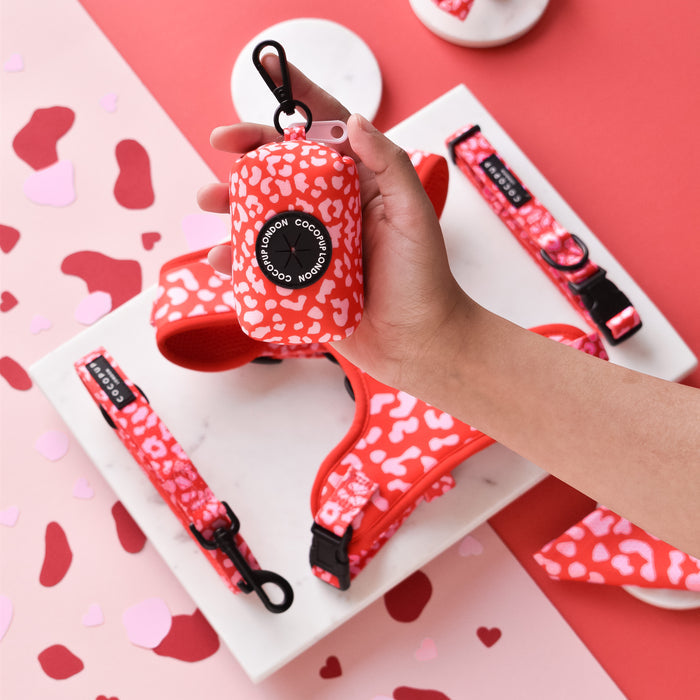 Red and pink leopard print poop bag holder by Cocopup London held in hand, with matching leash and harness shown.