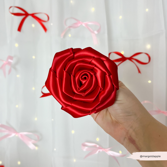 Hand holding a red rose collar flower in front of a white curtain with red and pink ribbon bows.
