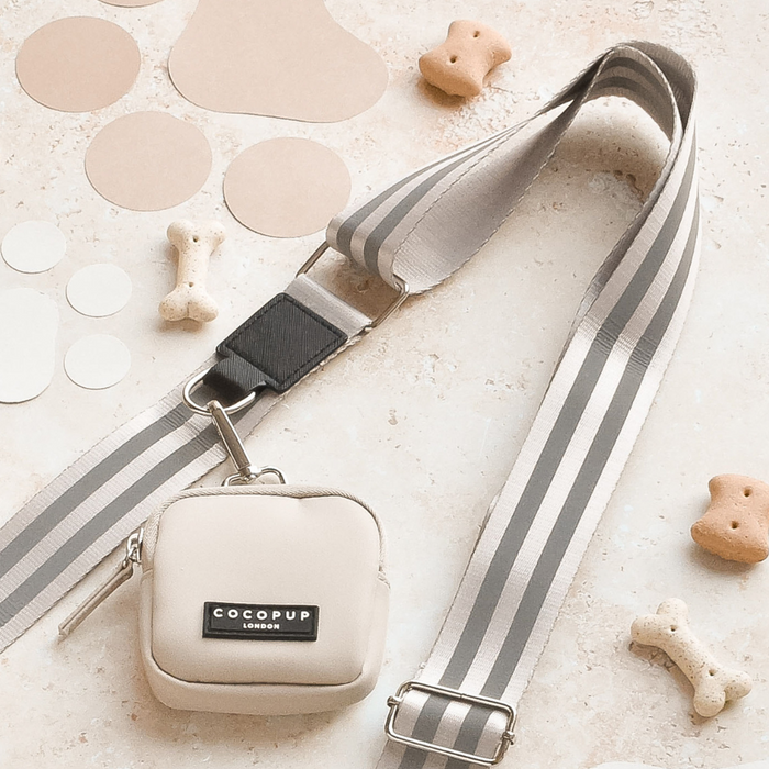 A stylish beige and gray striped dog leash is paired with the Reflective Bag Strap in Nude from Cocopup London, perfect for dog walking. The small beige carrying pouch, also labeled "Cocopup London," completes the set. Dog bone-shaped treats and round cutouts scattered around the leash and pouch on a light beige surface ensure both safety and style.