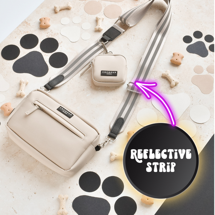 A beige crossbody bag with a small attached pouch and the Reflective Bag Strap - Nude from Cocopup London sit on a surface decorated with paw prints and dog bones. A bright arrow points to the strap, highlighting the "REFLECTIVE STRIP" text for added safety during dog walking.