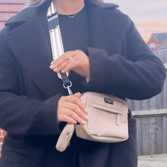 A person in a black coat adjusts a nude reflective Cocopup London bag strap on a beige crossbody bag outdoors.