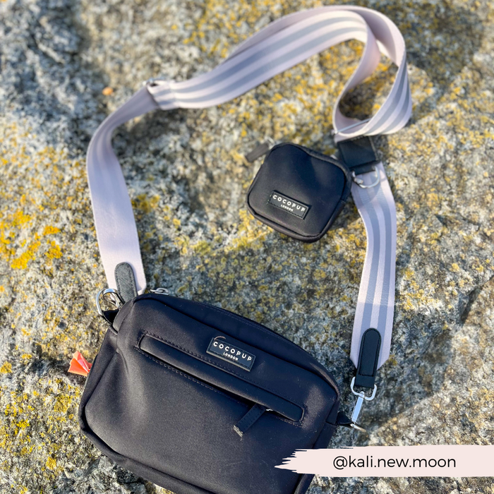 A black crossbody bag with a smaller pouch attached sits on a rocky surface. The bag and pouch prominently display the brand name "Cocopup London," and the strap is light purple with white stripes. Perfect for dog walking, the reflective strap adds an extra layer of safety. The Instagram handle @kali.new.moon is displayed in the bottom corner.