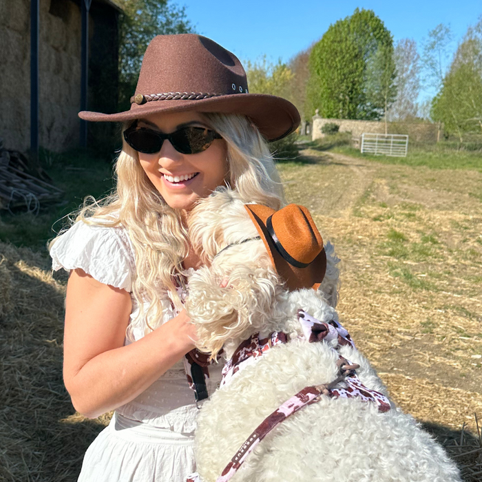 Woman in sunglasses hugs her dog wearing a brown Cocopup London Rodeo Pup Dog Hat, both smiling outside.