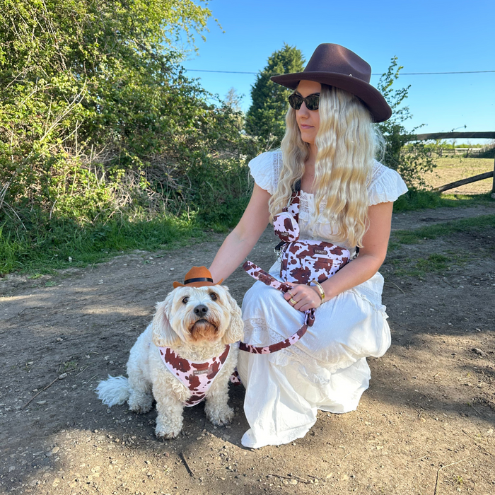 A woman in white kneels outside with her dog in a brown cowboy hat and matching bandana from Cocopup London.