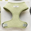 A green dog harness with heart patterns, named Luxe Adjustable Neck Harness - Sage Heart, lies on a flat surface next to a matching leash and other dog accessories. The harness features a gray buckle and a tag with the brand name "Cocopup London." The background showcases a light-colored textured surface.