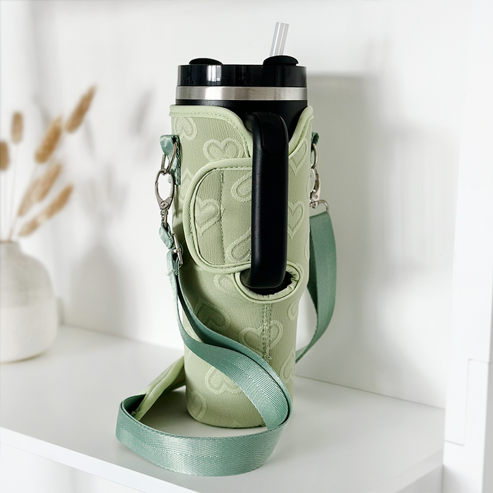 Sage green Stanley Quencher tumbler in a heart-patterned carry case, displayed on a white shelf.