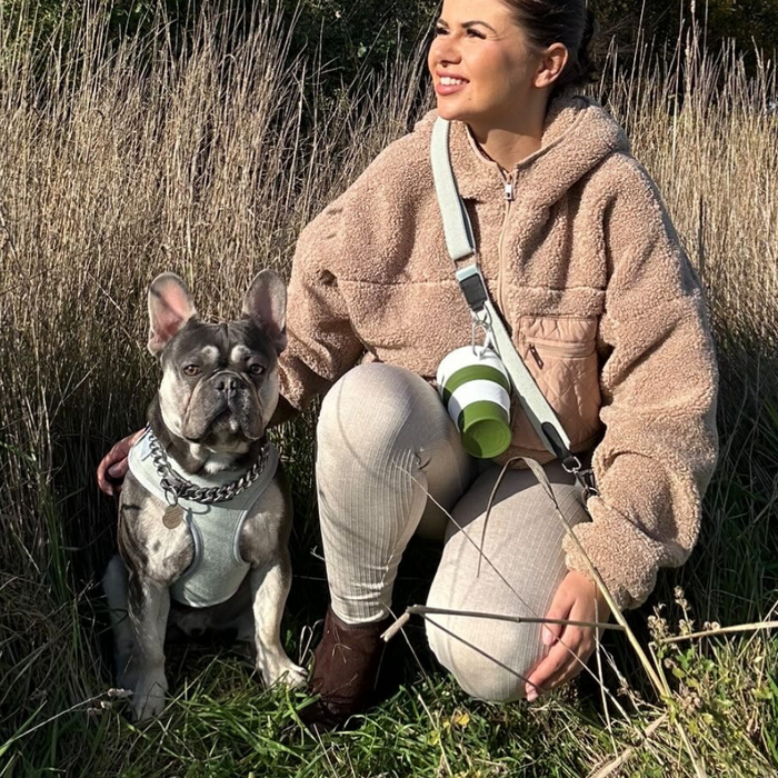 A person wearing a beige fleece jacket and light-colored pants is kneeling in tall grass next to a French Bulldog outfitted in the Cocopup London Sage Tweed Adjustable Neck Harness, Lead & Collar Bundle. The person is smiling and looking into the distance, with a plant holder and cup hanging from their shoulder.