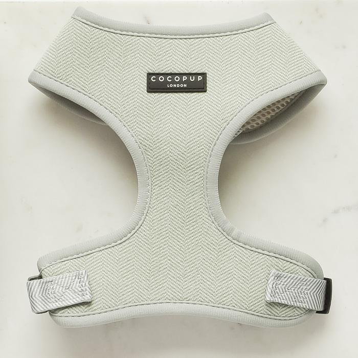 A light gray herringbone Sage Tweed Adjustable Neck Harness from Cocopup London is displayed on a white marble surface. This stylish dog harness features a black label with the brand name "Cocopup London" on the front, and is designed for both comfort and style with its fully adjustable straps and plastic buckles.