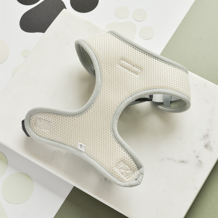 A light beige and gray Sage Tweed Adjustable Neck Harness from Cocopup London is placed on a white marble surface with a green and white background. The stylish dog harness, crafted from mesh material, appears to be comfortably padded. A small tag with the letter "S" indicates the size.