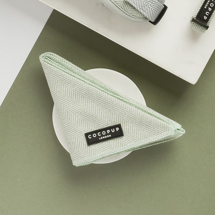 A green herringbone patterned pet bandana labeled "Cocopup London," known as the Sage Tweed Bandana, is neatly folded into a triangle and placed on a round white dish against a green background, showcasing its lightweight and breathable design. A matching country style pet accessory is visible at the top of the image.