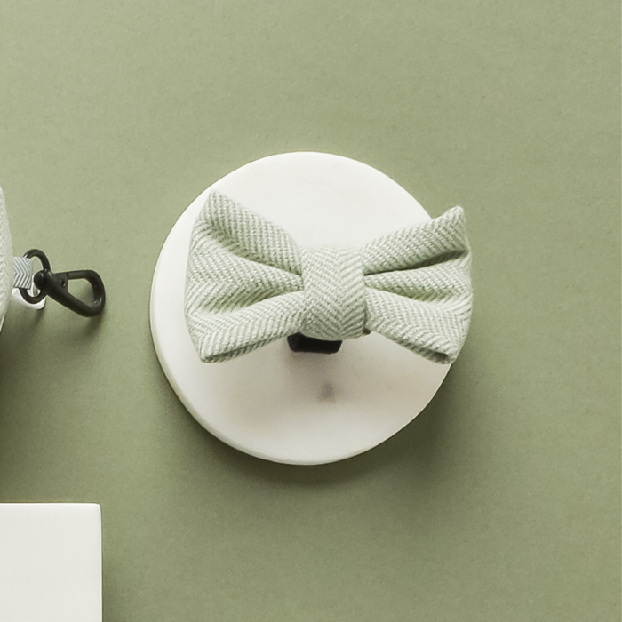 Sage green tweed dog bow tie with herringbone pattern displayed on a white stand against a green background.