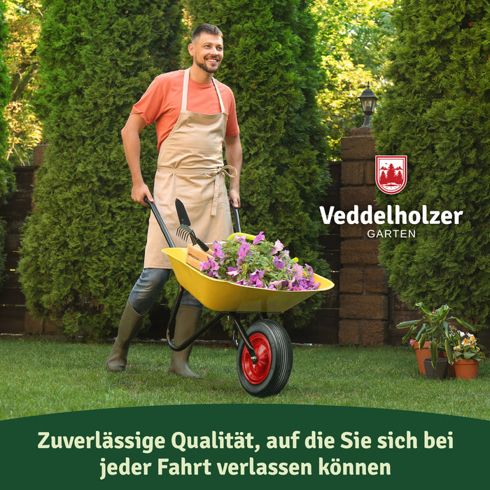 Mann mit Schürze und Gummistiefeln schiebt eine gelbe Schubkarre voller lila Blumen über den Rasen