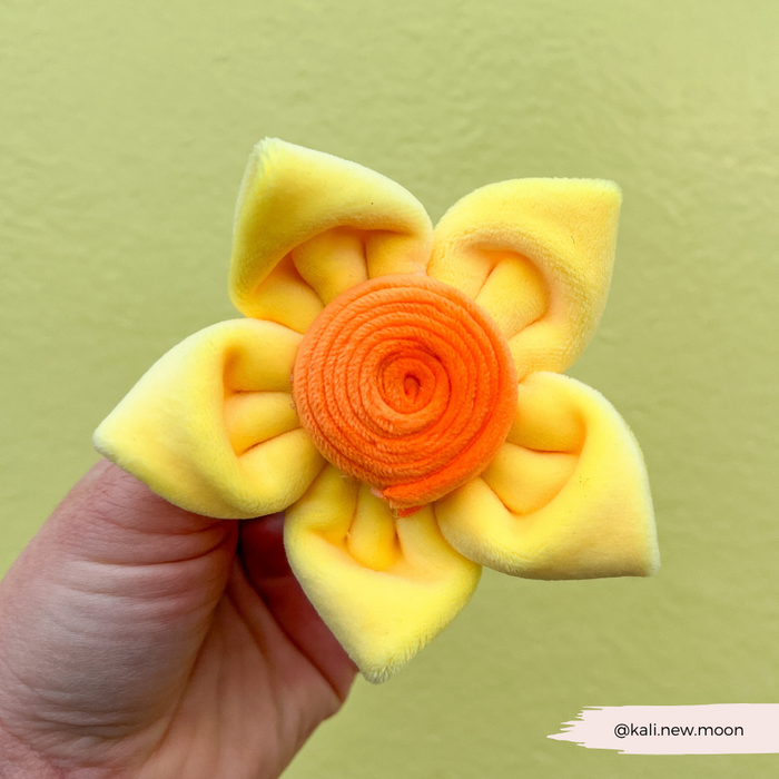 Hand holding yellow Cocopup London Spring Daffodil dog collar flower against a yellow background.