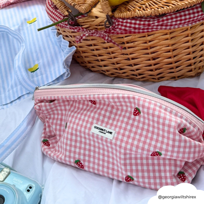 Pink expandable makeup bag with strawberry print beside picnic items on a white blanket.