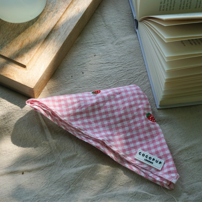 Pink strawberry-patterned dog bandana by Cocopup London, shown with an open book and wooden tray.