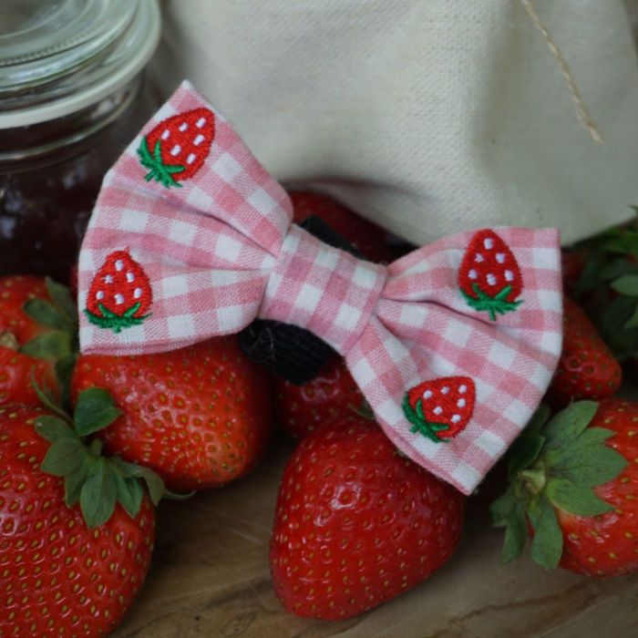 Pink gingham dog bow tie with embroidered strawberries, shown on a wooden surface.