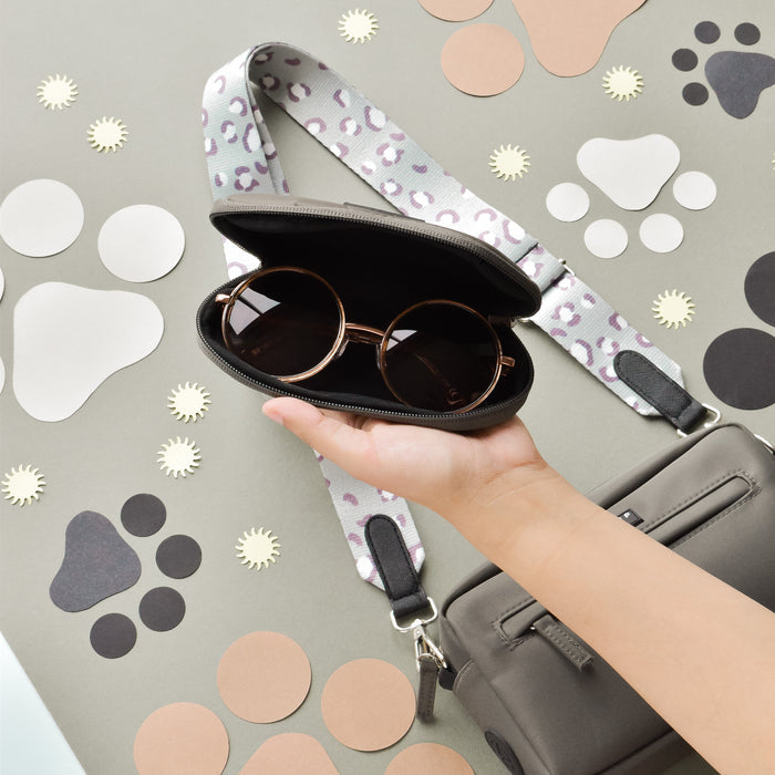 Hand holding a mocha Cocopup London sunglasses case with leopard strap next to a gray dog walking bag.