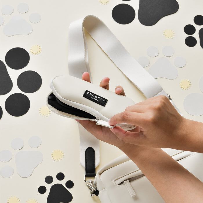 Hands unzip an oyster white Cocopup London sunglasses case; dog walking bag with black paw prints in the background.