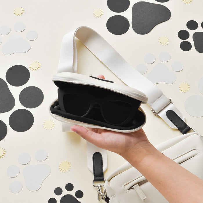 Hand holding a Cocopup London oyster white sunglasses case with black sunglasses, set against a paw print background.