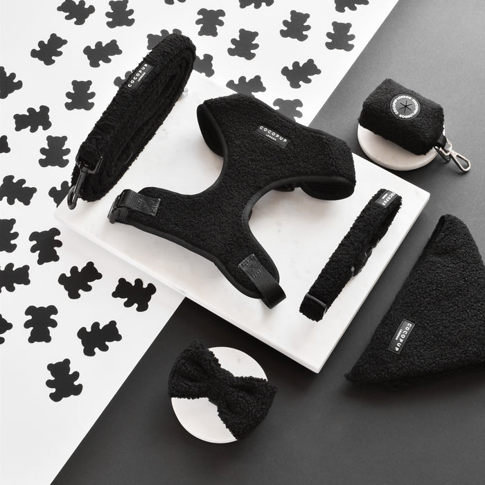 A collection of black dog accessories arranged on a white marble surface. Items include the Teddy Adjustable Neck Harness - Baloo by Cocopup London, leash, collar, waste bag holder, bandana, and bow tie, all featuring a teddy soft material. The background has a black-and-white pattern of bear silhouettes—perfect for your next dog walk.