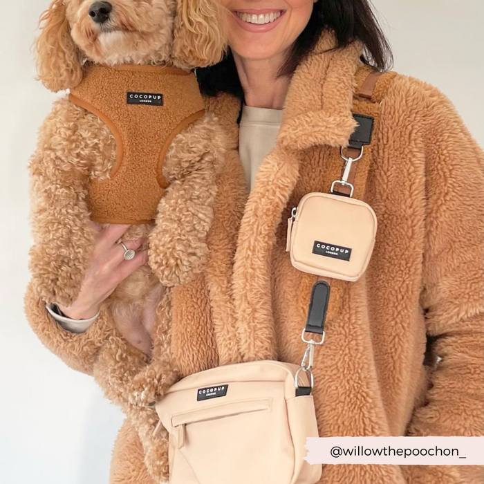 A woman in a brown fuzzy coat is holding a small dog wearing a matching Teddy Adjustable Neck Harness - Paddington from Cocopup London. She carries a beige crossbody bag and mini bag, perfectly completing her chic pupdrobe look. The user handle @willowthepoochon_ is displayed at the bottom right of the image.