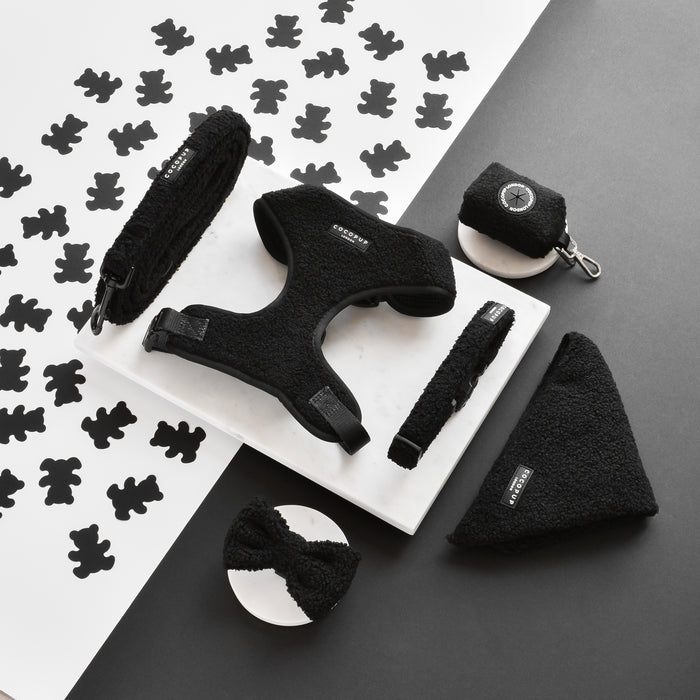 A black dog harness, leash, collar, bow tie, poop bag holder, and a Cocopup London Teddy Bandana - Baloo are artistically arranged on a white marble slab and a black-and-white background with teddy bear silhouettes. The accessories from the Cocopup London range of Bandanas are made from fluffy, fleecy material.
