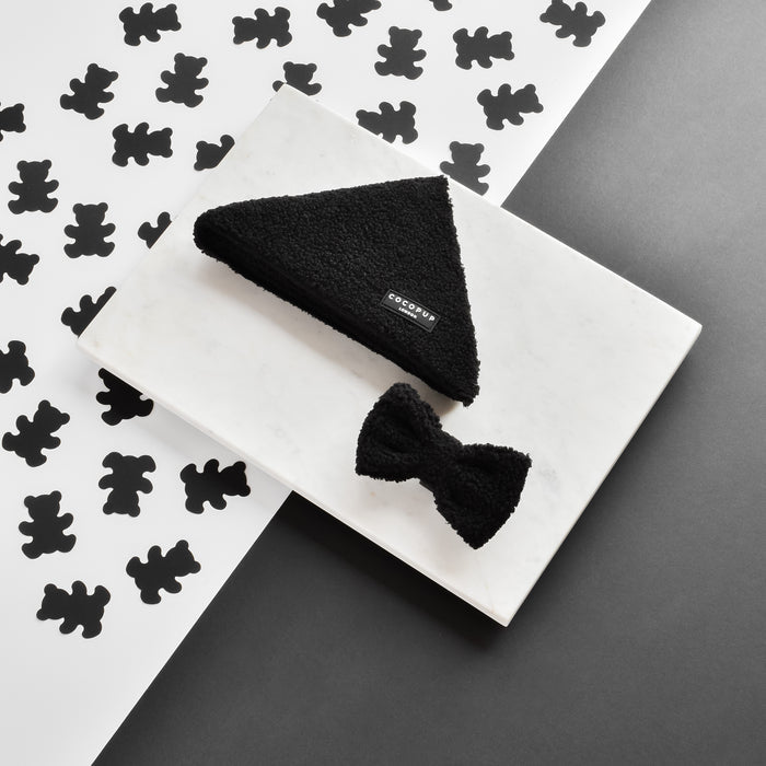 A Teddy Bandana - Baloo, a black triangular headscarf from Cocopup London, and a matching black bow tie are displayed on a white rectangular tray, set against a backdrop adorned with black bear silhouettes on white paper and plain black paper. The headscarf features a small rectangular label with text.
