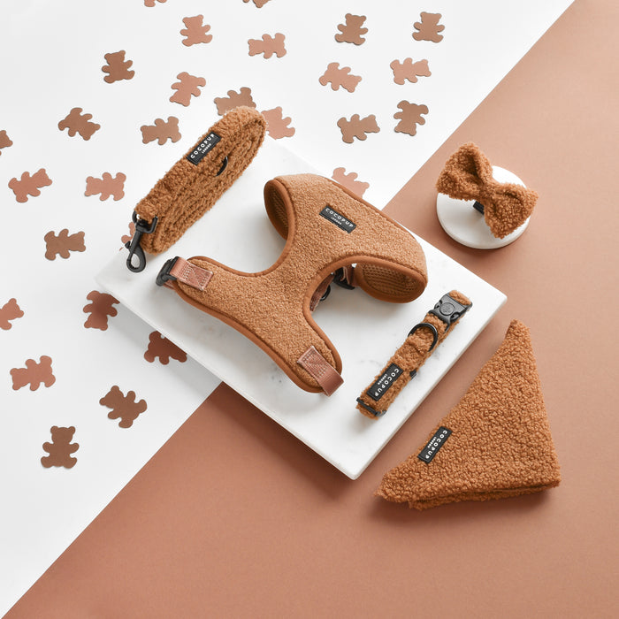 A flat-lay image showcases stylish brown dog accessories, including a harness, leash, bow tie, and two different types of collars from the Cocopup London range. Featured among them is the Teddy Bandana in Paddington design. These items are beautifully arranged on a marble and brown surface with small dog-shaped cutouts in the background.