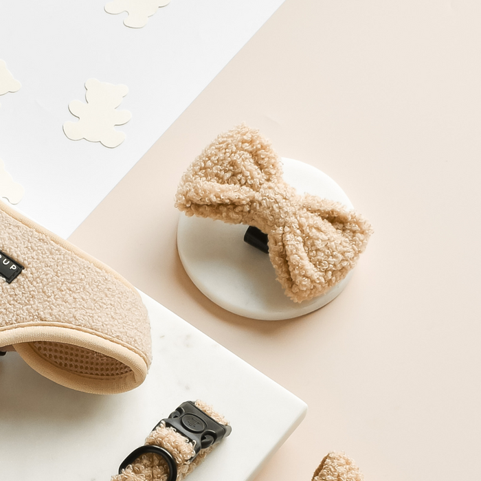 Tan textured Teddy Bow Tie by Cocopup London on a white stand, with matching tan dog accessories in the background.