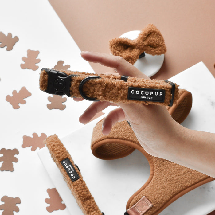 A hand holds a brown, fluffy dog collar featuring a black label with "Cocopup London." The same brand's Teddy Paddington Adjustable Neck Harness, Lead & Collar Bundle, which includes a neoprene harness, leash, and bow tie in brown tones, is displayed on a white marble surface surrounded by teddy bear-shaped cutouts.