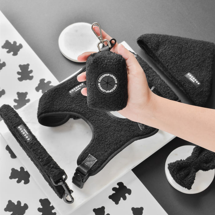 Hand holds a Cocopup London Teddy Poop Bag Holder in black, with matching black dog gear on a white background.