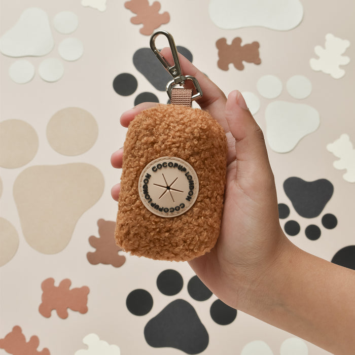 Hand holding brown Cocopup London Teddy Poop Bag Holder; paw and bone shapes in the background.