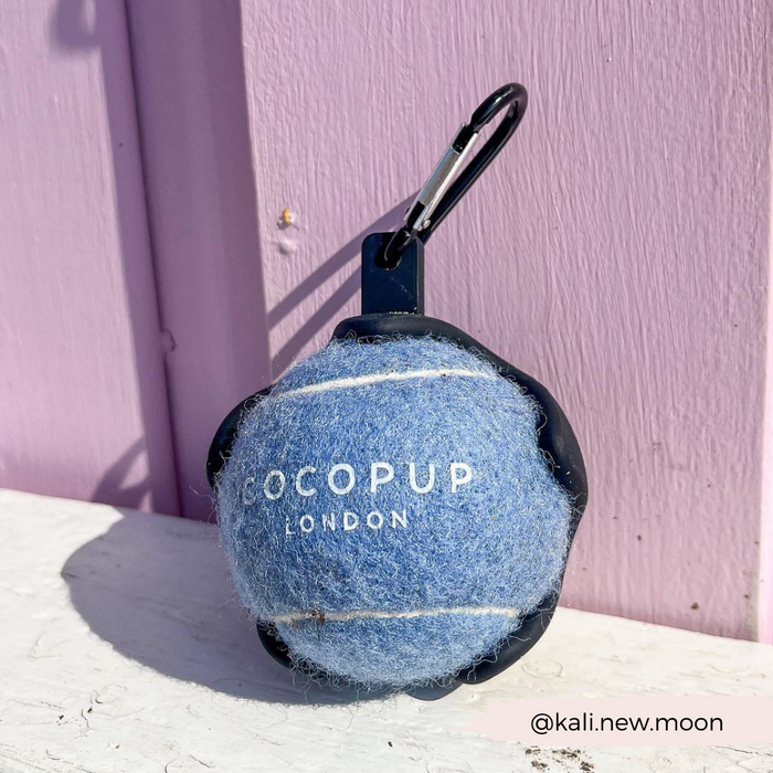 Blue tennis ball keychain with Cocopup London branding hanging on a hook against a pink wall.