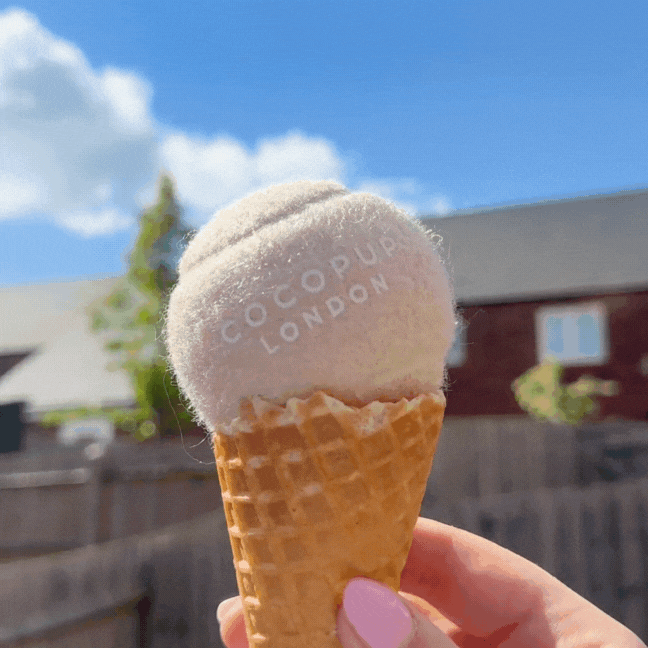 Hand holding a waffle cone topped with a tan Cocopup London tennis ball, resembling salted caramel ice cream.