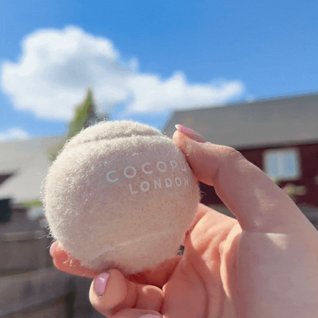 A hand holds three Cocopup London tennis balls in caramel, green, and pink against a clear blue sky.