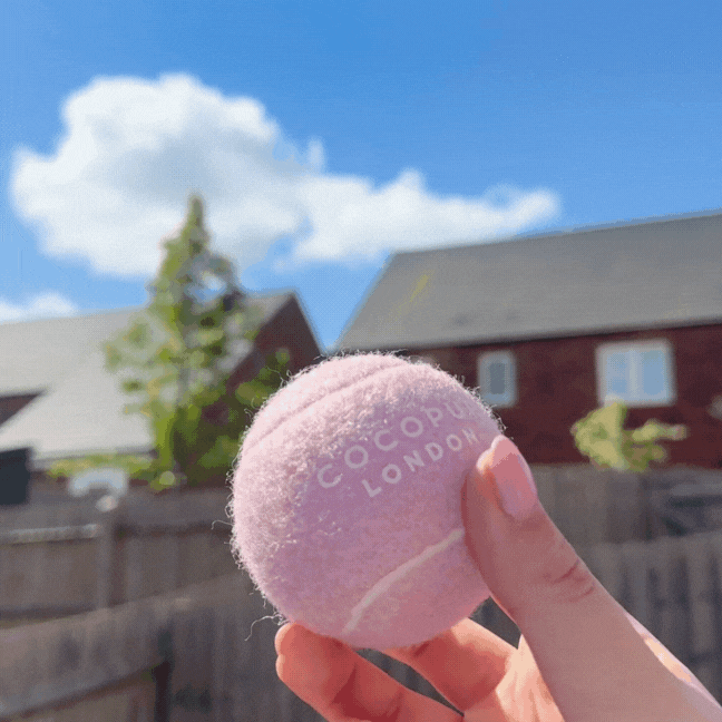 Hand holding a pink Cocopup London tennis ball outdoors, with houses visible in the background.