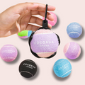 A hand holds a Cocopup London tennis ball holder with a tennis ball, surrounded by assorted colorful tennis balls.